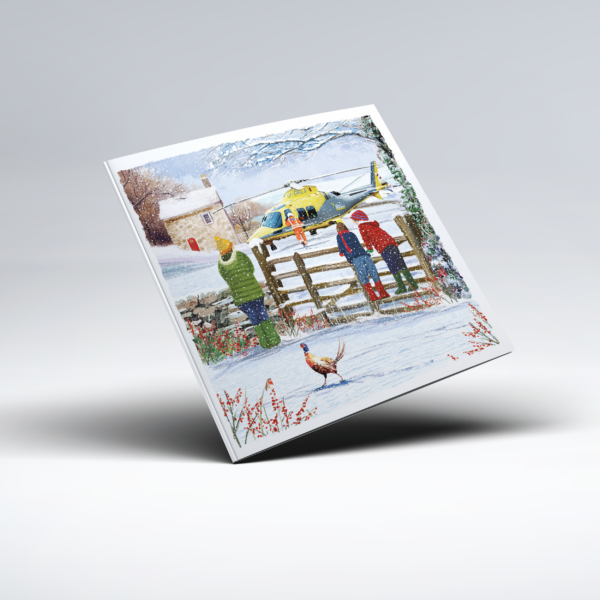 Illustrated Christmas card showing a yellow and grey Your Local Air Ambulance helicopter in a snow-covered countryside setting near a cottage and wooden gate. A mission crew member in an orange flight suit stands near the helicopter, while several people in colourful winter coats watch beside the gate; bare trees, snowy hills, a pheasant and red winter foliage complete the scene.