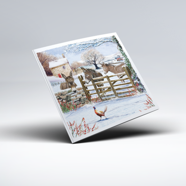Illustrated Christmas card showing two donkeys standing beside a snow-covered wooden gate, with a stone cottage, bare winter trees and snowy hills in the background. A pheasant stands in the foreground among red berries and colourful winter foliage, with snow covering the ground, trees and surrounding landscape.