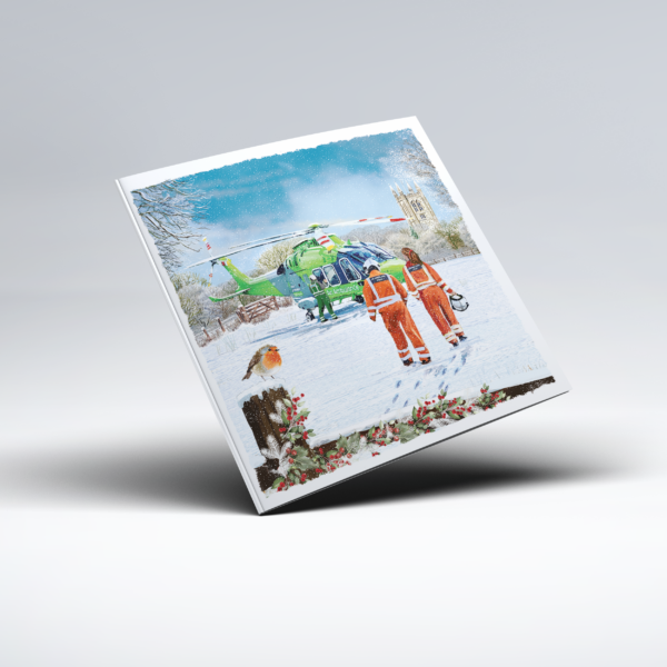 Illustrated Christmas card showing a green National Children’s Air Ambulance helicopter in a snow-covered rural setting. Three flight crew members are pictured near the helicopter, two wearing bright orange flight suits and one wearing green, while a robin perches in the snowy foreground; a wooden fence, bare winter trees, falling snow and a church tower complete the scene.