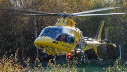 Yellow air ambulance helicopter hovering above grassy ground near trees, with spinning rotor blades visible