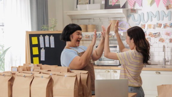 Two women celebrate success in a cheerful kitchen while packing paper bags, with a positive and energetic atmosphere.