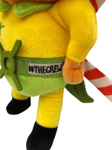 Photograph of stuffed bear plush, Blade - our TCAA mascot. He's wearing a yellow top, and a green cape and mask. On his back are helicopter blades. In this close up image, you can see his #TheCrew emblem on his waist.