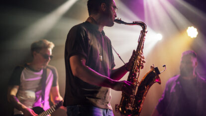 Saxophonist playing his saxophone during a live concert with his bandmates playing guitar and other instruments in the background under purple and orange stage lights